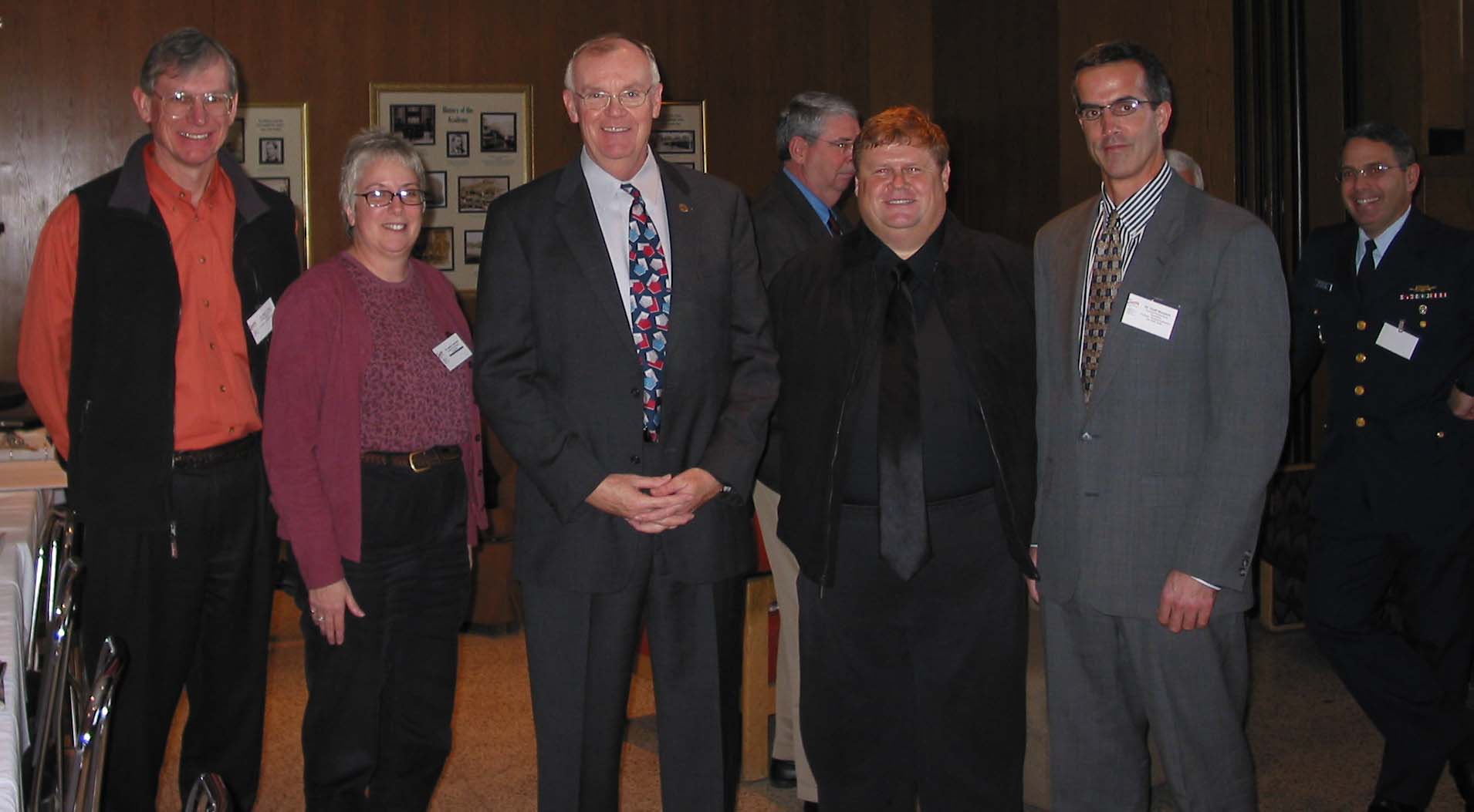A group of five people wearing name tags pose for a photo indoors, with another person standing in the background to the right. Framed pictures are displayed on the wall behind them.