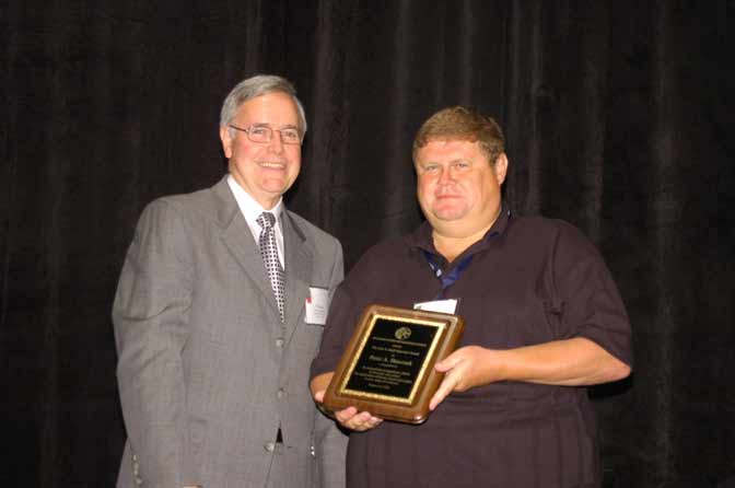 Two men are standing together, one in a suit and the other in a dark shirt, holding a plaque. The background is dark and plain.