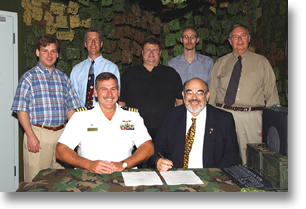 A group of seven men, including a uniformed officer and a man in a suit, pose around a table with documents and military decor in the background.