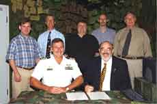 Seven men pose for a group photo, with two seated in front and five standing behind them. The seated men have documents in front of them, and one is in a military uniform.