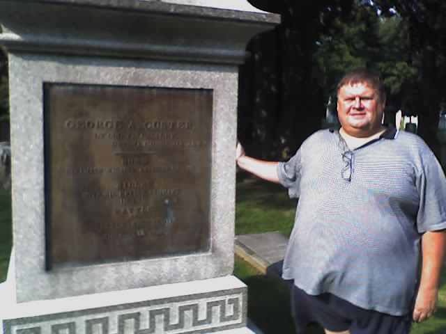 A man wearing a gray and black shirt stands beside a large stone monument in a cemetery.
