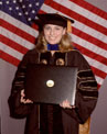 A person in academic regalia smiles and holds a diploma. An American flag is visible in the background.