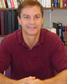 A person wearing a maroon shirt sits at a desk with bookshelves in the background.
