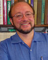 A person with glasses and a beard is smiling in front of a bookshelf while wearing a blue shirt.