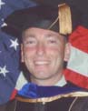 A smiling person in graduation attire stands in front of an American flag.
