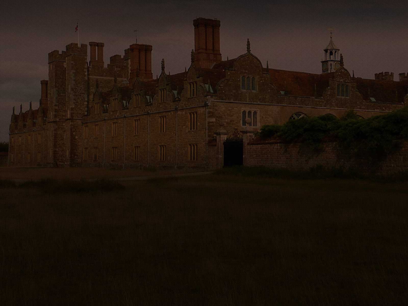 A historic stone building with multiple chimneys and gothic-style architecture stands against a dark, cloudy sky. The structure has tall windows and an archway, surrounded by grass and a stone wall.