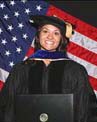 A smiling person in graduation attire stands in front of an American flag, holding a diploma.