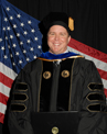 A person in academic regalia is smiling in front of an American flag.