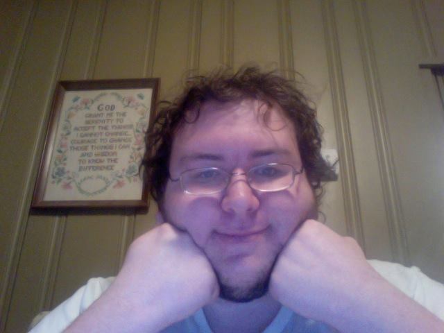 A person with glasses and a beard, resting their chin on their fists, smiles at the camera with a framed motivational quote in the background.