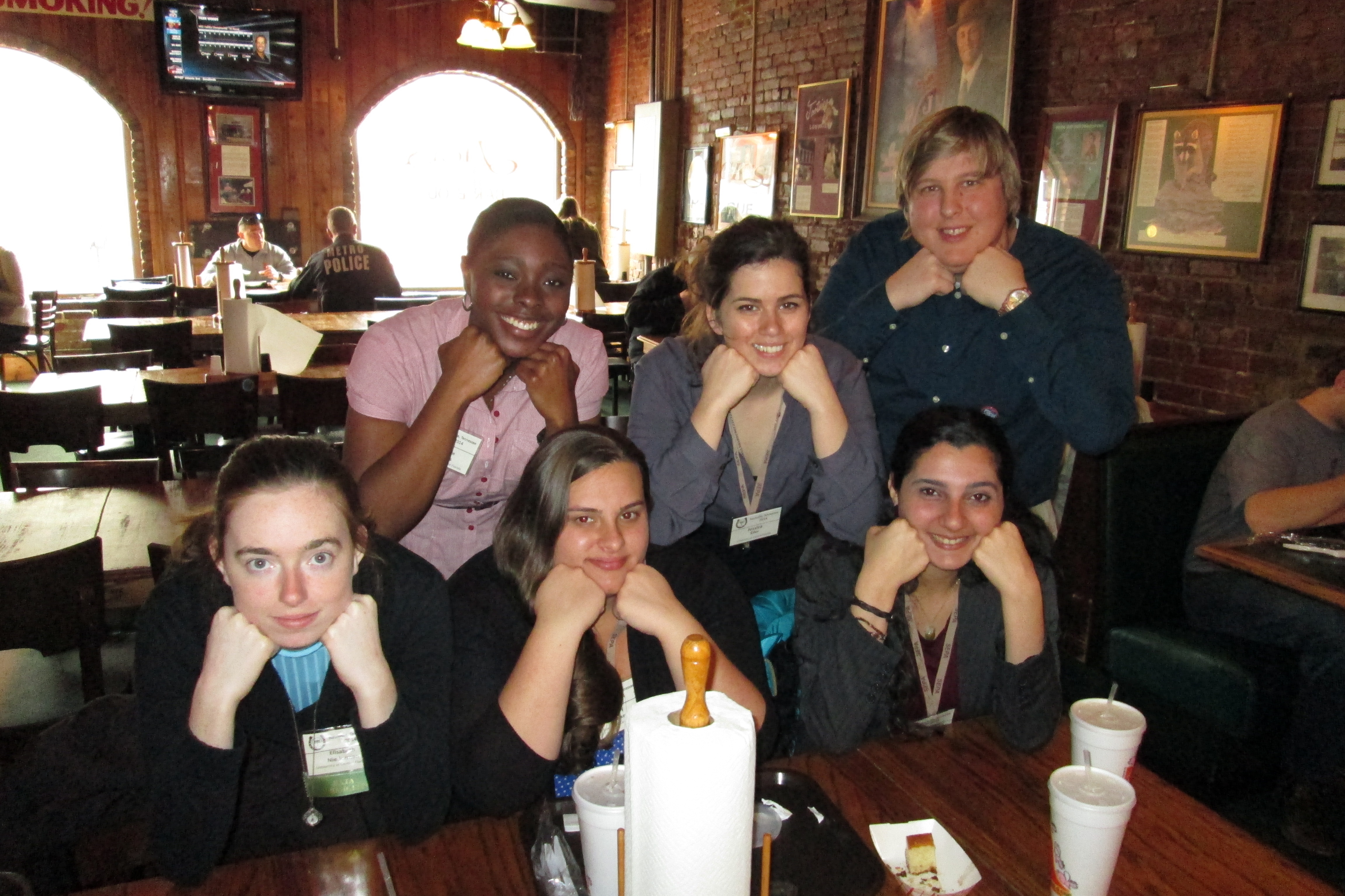 Six people sitting at a table in a restaurant, resting their chins on their hands and smiling at the camera. The background shows wooden walls, framed pictures, and other diners.