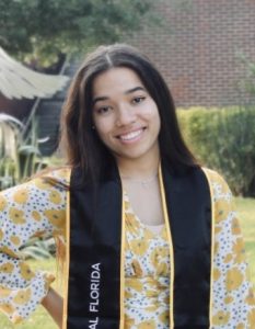 Young woman in a yellow floral dress wearing a graduation stole, standing outdoors and smiling at the camera.