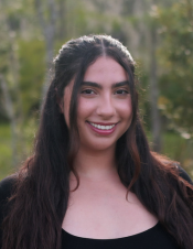 A woman with long dark hair and a black top smiles while standing outdoors with greenery in the background.